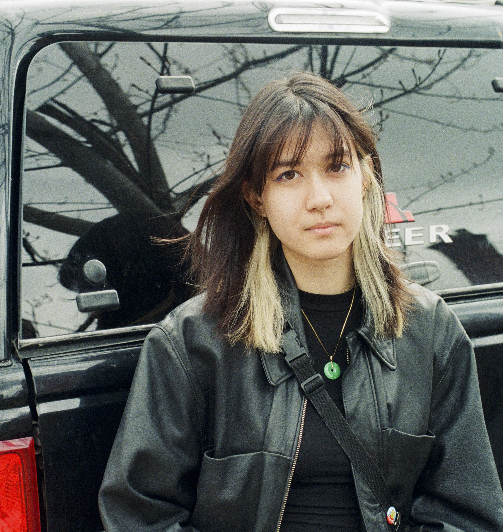 Image of person with brown and blonde hair, wearing black and a jade necklace, leaning against the back of a car.