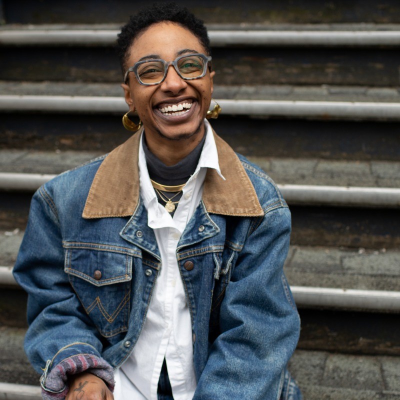 A brown-skinned non-binary person sits on a set of steps and smiles broadly, mid-laugh, facing the camera. Their black afro hair is cropped short with a fade. They have on square-shaped tortoise shell glasses and gold earrings. They also wear a jean jacket with a brown corduroy lapel. Underneath the jacket is a white button-up shirt with a grey turtleneck under that. Around their neck are a few gold necklaces. 